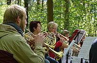 Mehrere Personen mit Blechbalsinstrumenten sitzen in einem Frühlingswald.