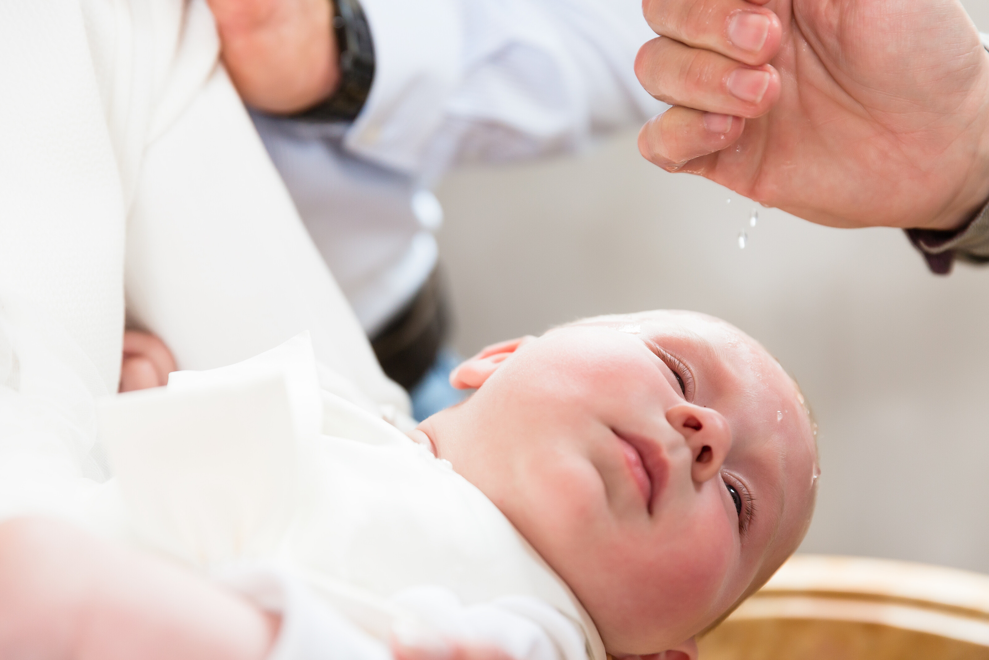Ein Baby wird über eine Taufschale gehalten und Wasser tropft auf seinen Kopf. In der kirchlichen Tradition findet die Taufe überwiegend im ersten Lebensjahr statt. 