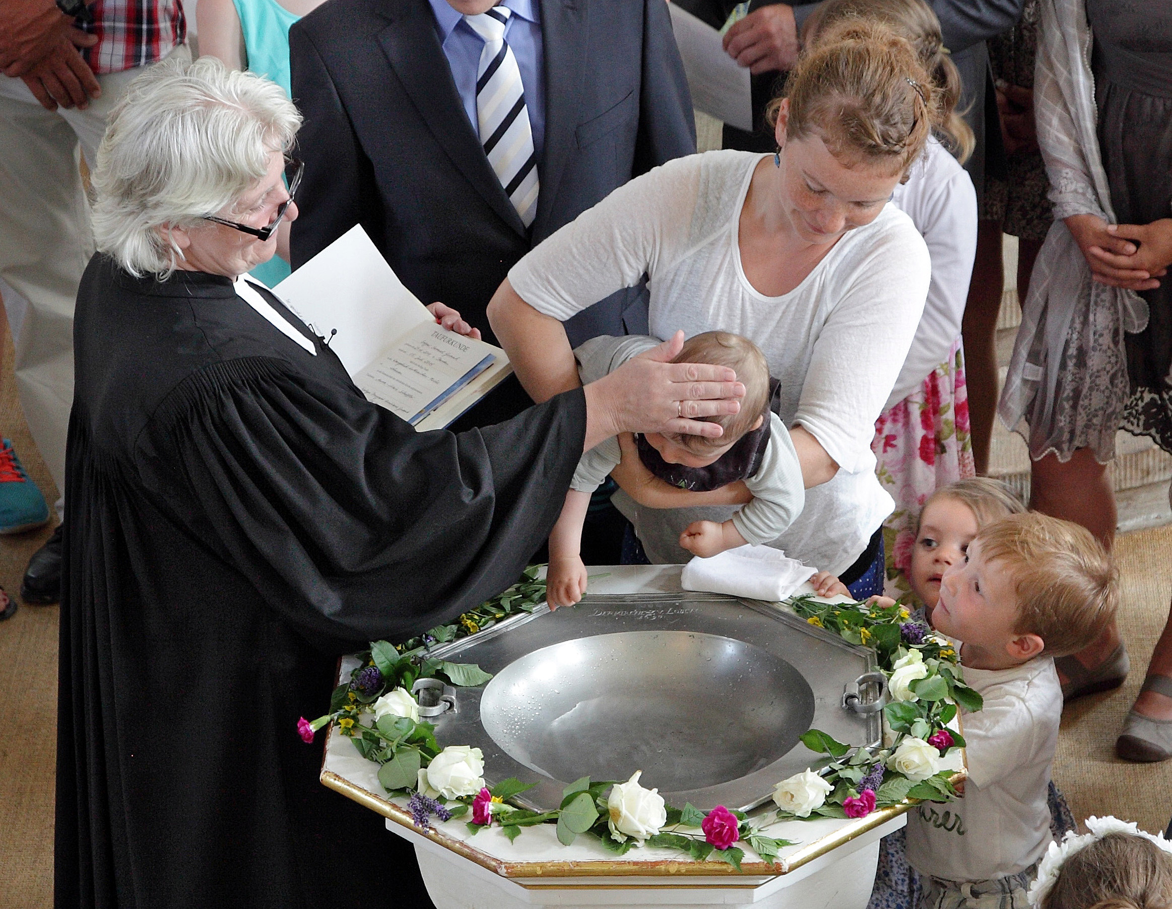 Eine Mutter hält ein Baby über die Taufschale, die Pfarrerin lässt Wasser über seinen Kopf rinnen. Die Taufe ist das Ritual zur Aufnahme in der christlichen Gemeinde. Sie ist einmalig und unwiederholbar. 