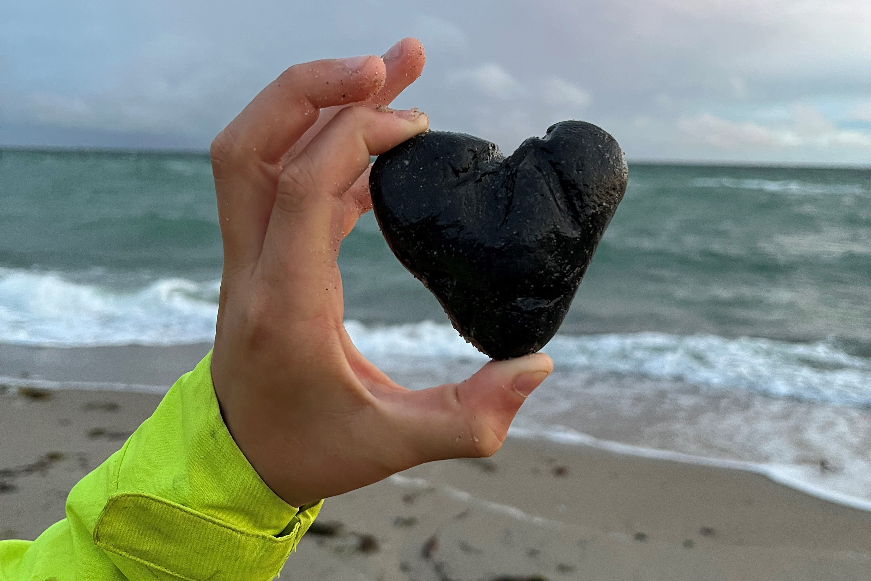 Hand hält herzförmigen schwarzen Stein, im Hintergrund Strand und Meer.